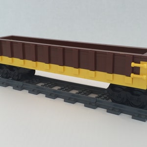 Puede incluir: Un vagón de tren LEGO marrón y amarillo con ruedas negras y un acoplamiento metálico. El vagón está sobre una vía LEGO gris.