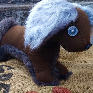 May include: A handmade brown plush toy horse with a gray mane and tail. The horse has button eyes and a button nose. The toy is sitting on a burlap sack.