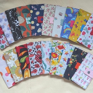 Puede incluir: Una colección de carteras rectangulares de tela dispuestas en dos filas, con varios patrones coloridos que incluyen globos aerostáticos, estampados florales, esqueletos, alimentos, arcoíris, fresas y diseños geométricos, cada una con un broche.
