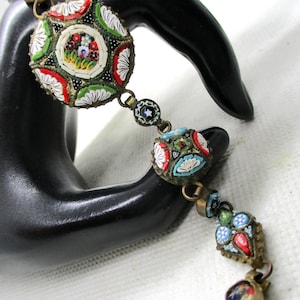 May include: A vintage mosaic bracelet with detailed patterns in red, white, green, and blue. The bracelet has a gold-toned metal setting and a clasp. The mosaic elements are round and square, displaying floral and geometric designs. The bracelet is a unique piece of jewelry.