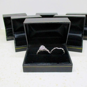 May include: An open black ring box with a diamond engagement ring and a diamond wedding band. The ring box is lined with black velvet and has a gold trim. Several other closed ring boxes are in the background.