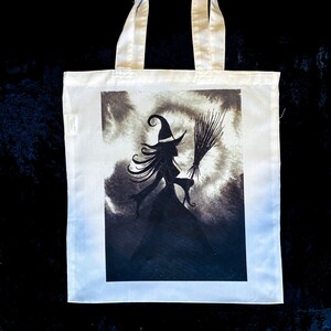 Pu&ograve; includere: Borsa tote bianca con un'illustrazione in bianco e nero di una strega con una scopa in un cielo nuvoloso.