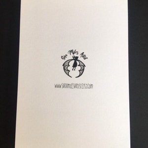 May include: A white card with a black bat logo and the text "Sara Miles Artist Etsy.com". The logo features a bat with a crescent moon and stars. The card is on a black surface.