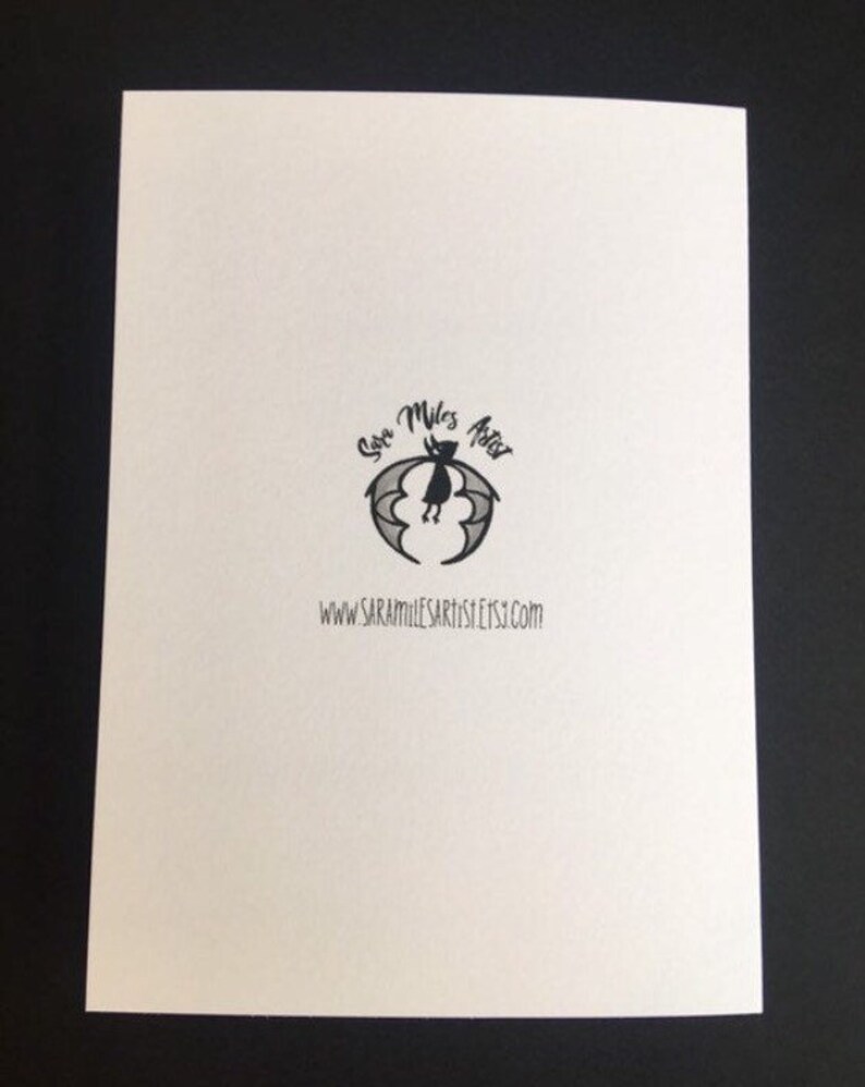 May include: A white card with a black bat logo and the text "Sara Miles Artist Etsy.com". The logo features a bat with a crescent moon and stars. The card is on a black surface.