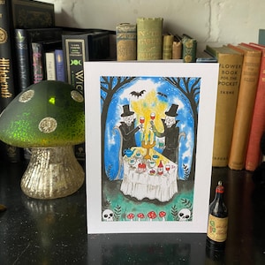 May include: A watercolour illustration shows two skeleton figures in top hats toasting with wine glasses at a table set with food and drinks. The scene is framed by trees, bats, and a night sky. A small bottle of wine is in the foreground.