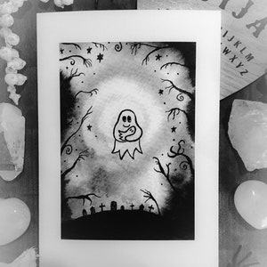 May include: Black and white artwork of a ghost holding a smaller ghost, set in a spooky forest scene with a graveyard. The artwork is framed in white, with a Ouija board and decorative crystals in the background.