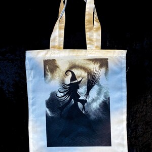 Pu&ograve; includere: Una borsa tote bianca con una silhouette nera di una strega che vola su una scopa su uno sfondo di cielo tempestoso.
