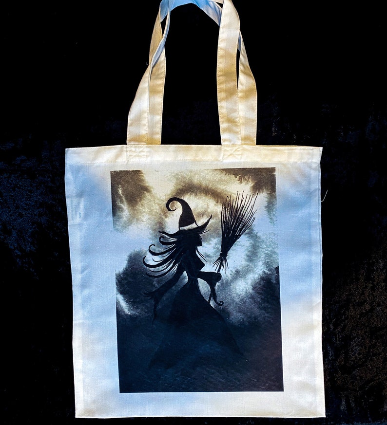 Pu&ograve; includere: Una borsa tote bianca con una silhouette nera di una strega che vola su una scopa su uno sfondo di cielo tempestoso.