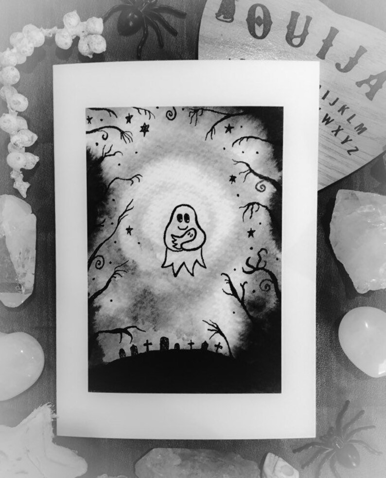 May include: Black and white artwork of a ghost holding a smaller ghost, set in a spooky forest scene with a graveyard. The artwork is framed in white, with a Ouija board and decorative crystals in the background.