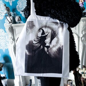 Pu&ograve; includere: Borsa tote bianca con un'illustrazione in bianco e nero di una strega che vola su una scopa con un gatto. La strega indossa un cappello a punta e ha lunghi capelli fluenti.