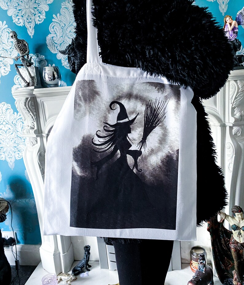 Pu&ograve; includere: Borsa tote bianca con un'illustrazione in bianco e nero di una strega che vola su una scopa con un gatto. La strega indossa un cappello a punta e ha lunghi capelli fluenti.