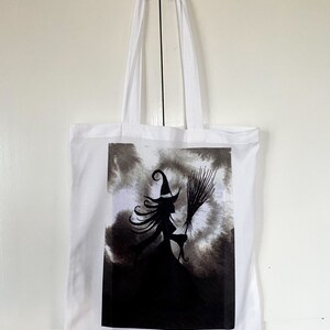 Pu&ograve; includere: Borsa tote bianca con un'illustrazione in bianco e nero di una strega che vola su una scopa su uno sfondo di nuvole scure.