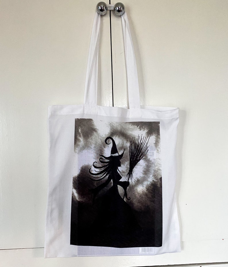 Pu&ograve; includere: Borsa tote bianca con un'illustrazione in bianco e nero di una strega che vola su una scopa su uno sfondo di nuvole scure.