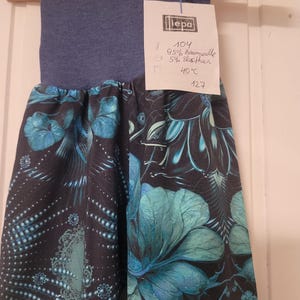 Puede incluir: Un pantal&oacute;n con una cintura ancha azul y una tela negra con un estampado floral en tonos turquesa y azul. Tiene una etiqueta blanca adjunta con el texto "iepa", "104", "95% Baumwolle", "5% elasthan", "40&deg;C" y "127" escrito a mano.