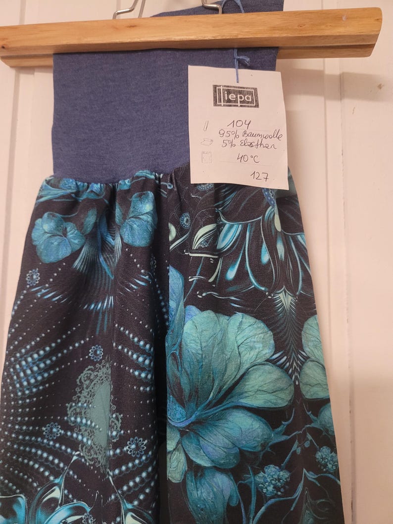 Puede incluir: Un pantal&oacute;n con una cintura ancha azul y una tela negra con un estampado floral en tonos turquesa y azul. Tiene una etiqueta blanca adjunta con el texto "iepa", "104", "95% Baumwolle", "5% elasthan", "40&deg;C" y "127" escrito a mano.