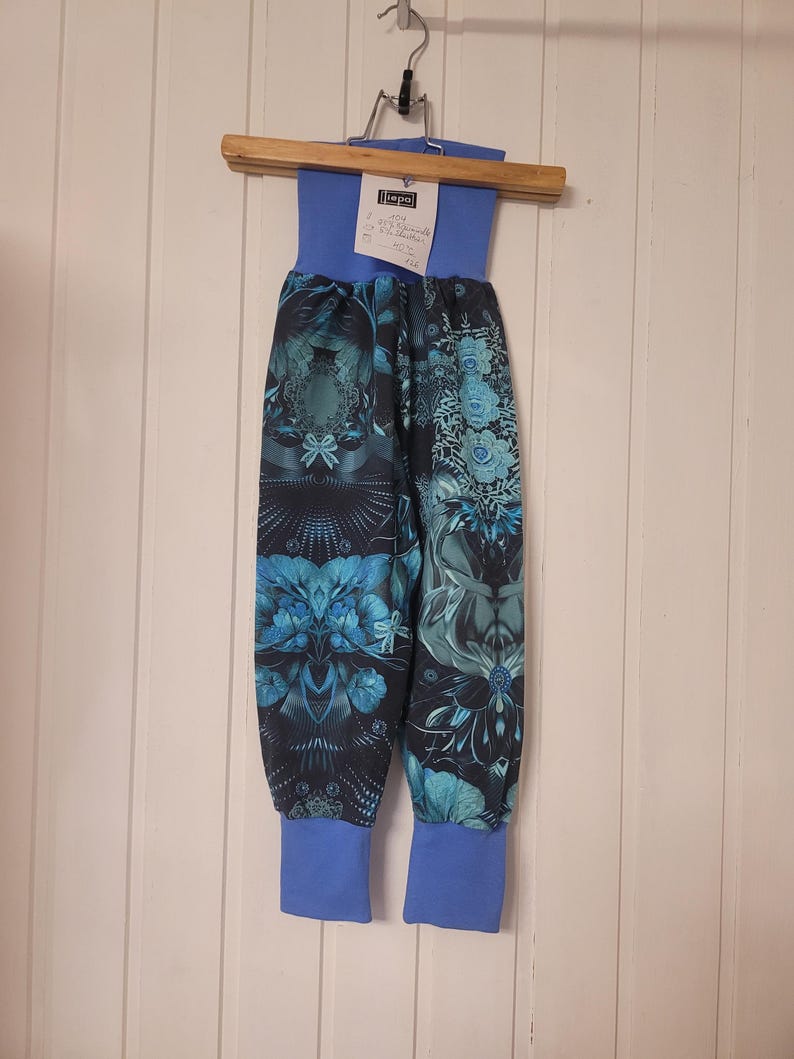 Puede incluir: Pantalones azules con un patr&oacute;n floral y geom&eacute;trico oscuro e intrincado en tonos verde azulado y negro. La prenda tiene una cintura el&aacute;stica ancha de color azul s&oacute;lido y pu&ntilde;os acanalados azules a juego en los tobillos. Una etiqueta blanca con texto est&aacute; sujeta a la cintura.