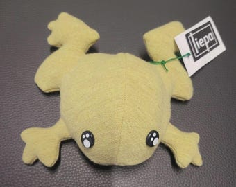 Puede incluir: Un juguete de peluche con forma de rana de color amarillo claro con ojos de botón blancos y negros y cuatro patas palmeadas. Una etiqueta blanca con la palabra "iepa" está sujeta con un hilo verde. La rana está sobre una superficie gris oscuro.