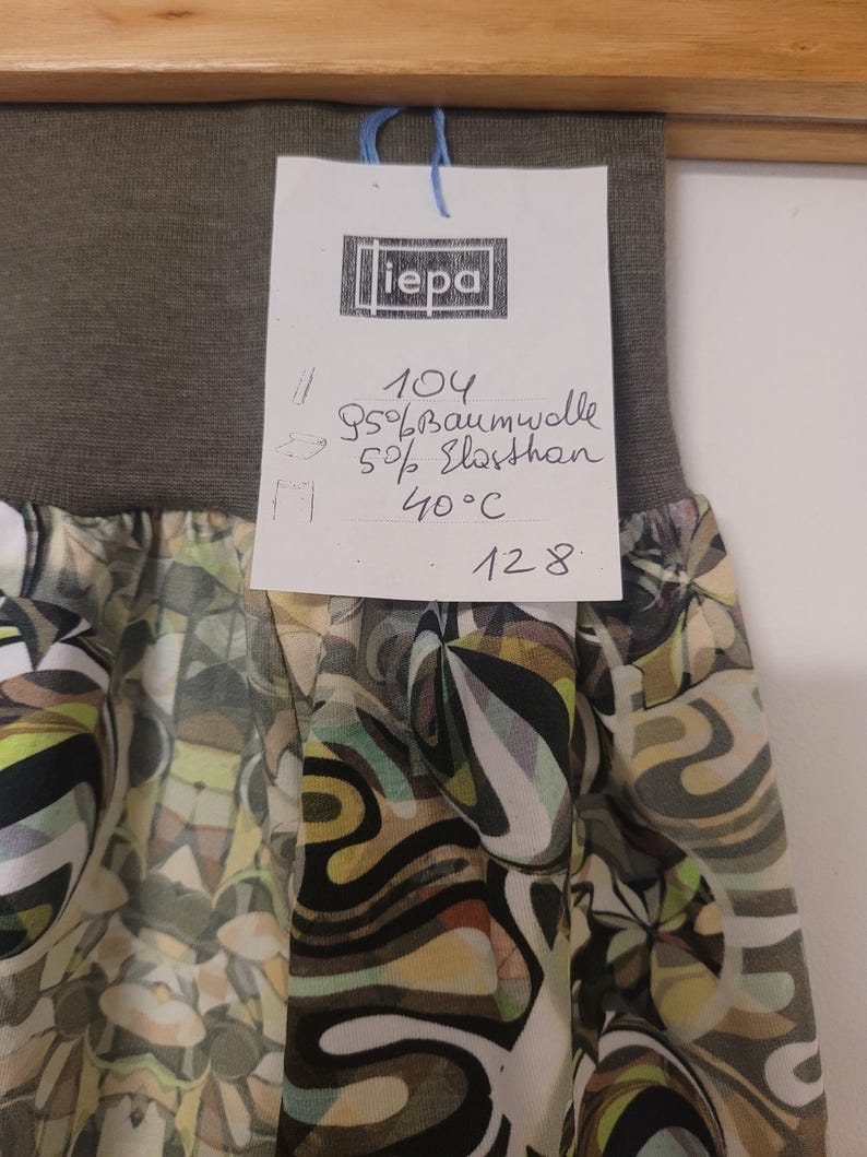 Puede incluir: Una falda con una cintura el&aacute;stica ancha de color verde oliva oscuro y una secci&oacute;n inferior estampada con formas abstractas en tonos verde oliva, negro, blanco y amarillo p&aacute;lido. Una etiqueta blanca muestra el texto iepa, 104, 95% Baumwolle, 5% Elasthan, 40&deg;C y 128.