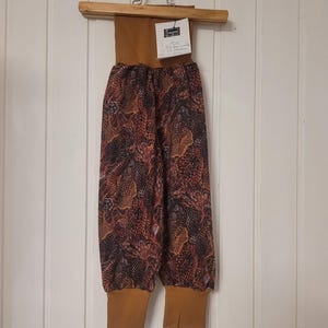 Puede incluir: Pantalones con una cintura alta de canal&eacute; color mostaza y pu&ntilde;os a juego en los tobillos. La tela principal presenta un patr&oacute;n oscuro e intrincado de formas abstractas en naranja, marr&oacute;n y negro. Se incluye una etiqueta blanca con el texto "liepa".