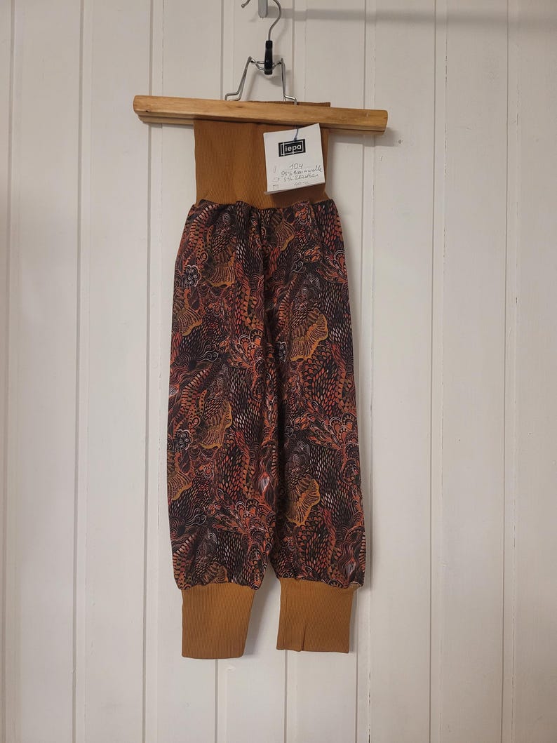 Puede incluir: Pantalones con una cintura alta de canal&eacute; color mostaza y pu&ntilde;os a juego en los tobillos. La tela principal presenta un patr&oacute;n oscuro e intrincado de formas abstractas en naranja, marr&oacute;n y negro. Se incluye una etiqueta blanca con el texto "liepa".