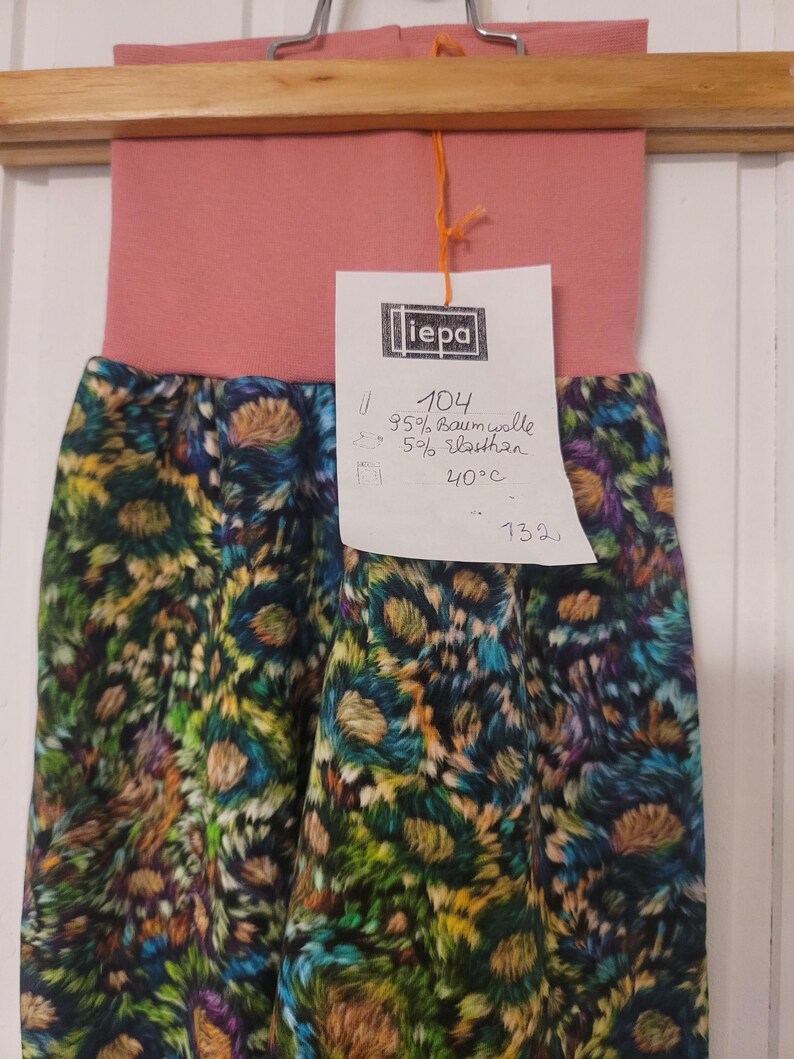 Puede incluir: Un pantal&oacute;n con una cintura ancha de color rosa empolvado y un estampado floral multicolor con tonos verdes, azules y amarillos. Una etiqueta blanca sujeta con un cordel naranja muestra el texto iepa, 104, 95% Baumwolle, 5% Elasthan, 40&deg;C y 132.