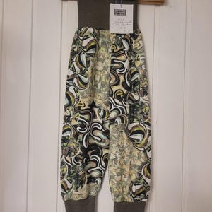 Puede incluir: Pantalones con una cintura alta de color marr&oacute;n oscuro s&oacute;lido y pu&ntilde;os a juego en los tobillos. La tela presenta un patr&oacute;n abstracto multicolor con formas arremolinadas en negro, blanco, verde oliva y amarillo. Tiene una etiqueta blanca.