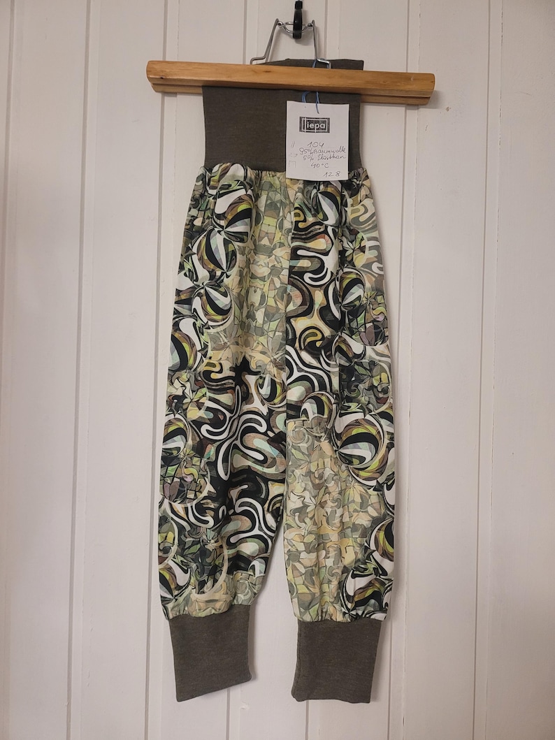 Puede incluir: Pantalones con una cintura alta de color marr&oacute;n oscuro s&oacute;lido y pu&ntilde;os a juego en los tobillos. La tela presenta un patr&oacute;n abstracto multicolor con formas arremolinadas en negro, blanco, verde oliva y amarillo. Tiene una etiqueta blanca.