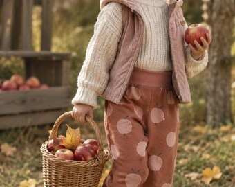 Puede incluir: Un niño o niña viste un suéter de punto color crema, un chaleco con capucha rosa polvoriento y pantalones de chándal color óxido con estampado de manzanas. Sostiene una cesta de mimbre llena de manzanas rojas y una manzana en la mano.