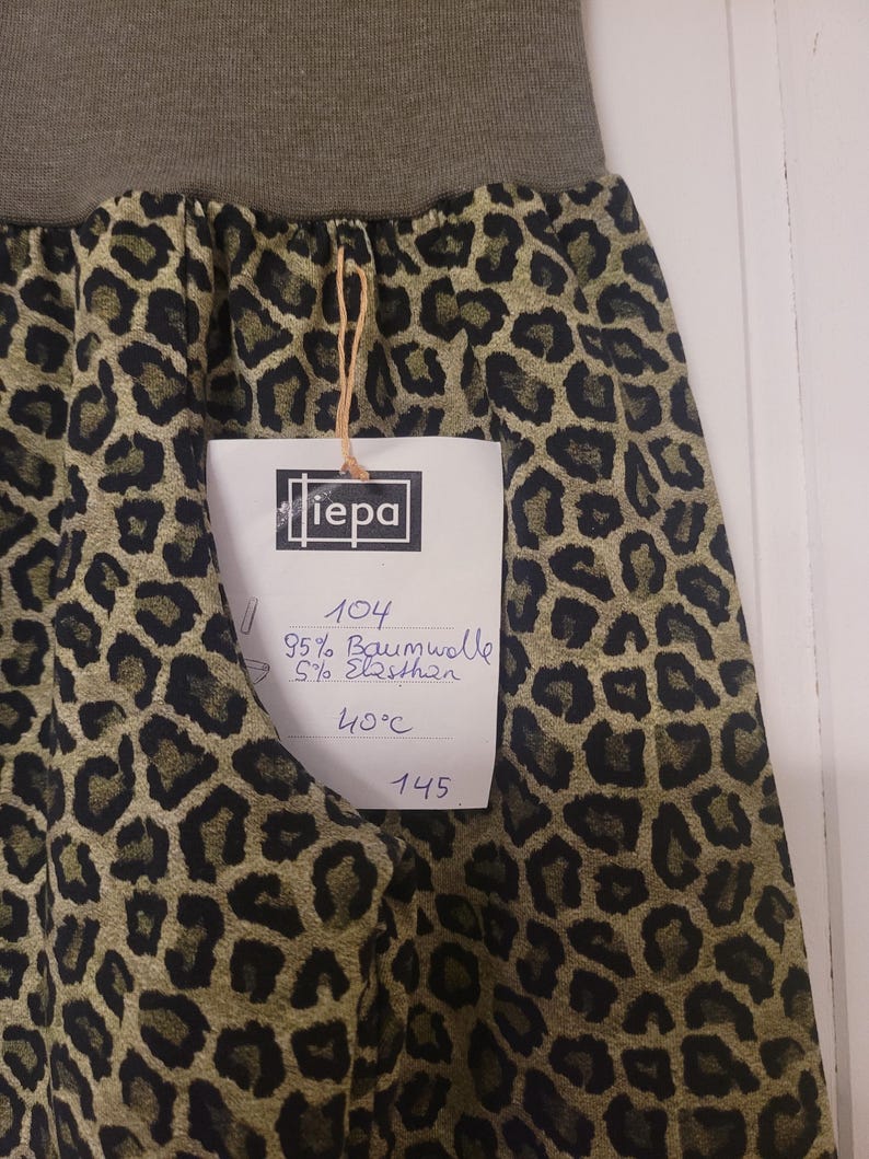 Puede incluir: Una falda con estampado de leopardo en verde oliva y negro con una cintura el&aacute;stica en verde oliva s&oacute;lido. Una etiqueta blanca unida con un cordel muestra el texto "iepa", "104", "95% Baumwolle", "5% Elasthan", "40&deg;C" y "145".