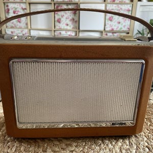 May include: A portable Bush radio featuring a textured brown plastic casing and a silver metal speaker grille. The device includes a brown carrying handle attached to the top, along with silver control knobs and buttons positioned on the upper panel.