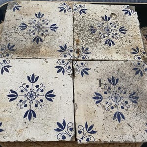 May include: Four square ceramic tiles arranged in a grid, each featuring a weathered off-white surface with a central dark blue floral motif and matching corner accents. The tiles show signs of age with visible surface wear and texture.