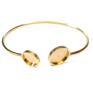 May include: A gold-colored open cuff bracelet with two oval settings. The bracelet is made of a thin, curved metal band, with the settings at each end. The settings have a shallow, recessed area.