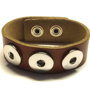 Könnte beinhalten: Ein braunes Lederarmband mit vier silbernen Druckknöpfen. Das Armband ist verstellbar und hat ein schlichtes Design.