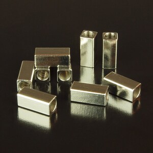May include: A group of silver metal end caps for cords or cables. The end caps are square shaped with a hole in the center.