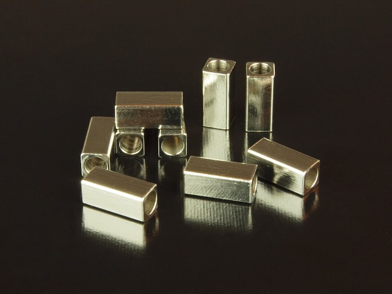 May include: A group of silver metal end caps for cords or cables. The end caps are square shaped with a hole in the center.