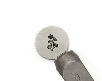 May include: A close-up of a metal stamp featuring a design of a small plant with leaves. The stamp is being used to create an impression on a metal disc.
