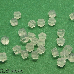 Op de afbeelding: Een close-up van een groep van 20 doorzichtige plastic oorbelruggen. De ruggen zijn gevormd als een bloem met vier bloemblaadjes. De ruggen zijn 4 mm bij 2,5 mm groot. De ruggen liggen op een groene ondergrond.