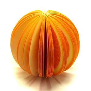 Op de afbeelding: Oranje kleverige notitieblok in de vorm van een bol met een gesegmenteerd ontwerp. Het blok is gemaakt van meerdere lagen papier, elk met een andere oranje tint.