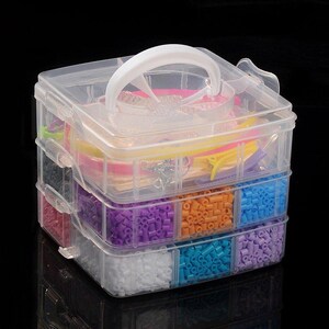 Può includere: Un organizer per artigianato in plastica trasparente con manico bianco. L'organizer ha tre scomparti impilati pieni di perline colorate e forniture per l'artigianato. Le perline sono di vari colori, tra cui viola, arancione, blu e bianco.