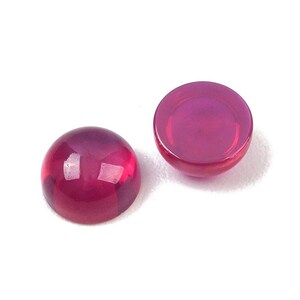 Könnte beinhalten: Zwei Cabochon-Edelsteine in tiefem Magenta. Die Edelsteine sind rund mit einer glatten, polierten Oberfläche, die das Licht reflektiert. Die Edelsteine sind vor einem weißen Hintergrund platziert.