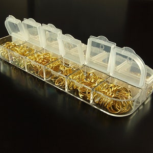 Op de afbeelding: Een transparante plastic opbergdoos met meerdere compartimenten gevuld met gouden springringen. De doos heeft een scharnierend deksel met een transparant plastic venster.