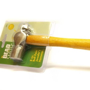 Könnte beinhalten: Ein 8 oz Ball-Pein-Juwelierhammer mit einem Holzgriff. Der Hammer ist in einer durchsichtigen Plastiktüte mit einem grün-weißen Etikett verpackt, auf dem "The Bead Smith" steht.