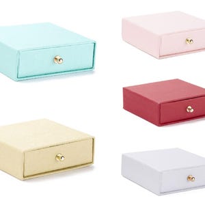Könnte beinhalten: Eine Sammlung kleiner, quadratischer Schmuckkästchen in verschiedenen Farben, darunter Hellblau, Rosa, Rot, Beige und Weiß. Jede Box hat eine Schublade mit einem kleinen, goldfarbenen Knopf. Die Boxen stehen vor weißem Hintergrund.