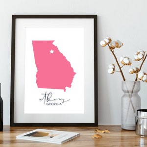 May include: A framed print of a pink outline of the state of Georgia with a white star marking the location of Athens, Georgia. The text "athens GEORGIA" is written below the map.