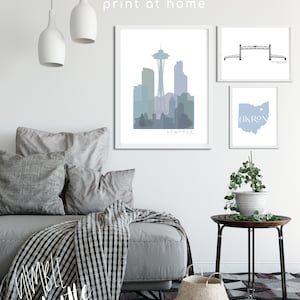 May include: Three printable wall art prints featuring a skyline of Seattle, a bridge, and the state of Ohio with the word "Akron" on it. The prints are framed in white and are displayed on a white wall with a gray couch and a side table with a plant.