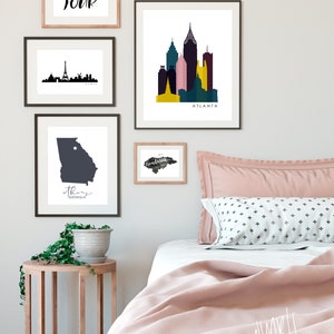 May include: A bedroom interior with a pink bedspread and a white headboard. There are several framed prints on the wall, including a black and white silhouette of the Paris skyline, a colorful illustration of the Atlanta skyline, and a gray outline of the state of Georgia. The text "BON! TOUR" is written in black on a white background in one of the frames. The text "Atlanta" is written in black on a white background in another frame. The text "Georgia" is written in black on a white background in another frame. The text "Honduras" is written in black on a white background in another frame.