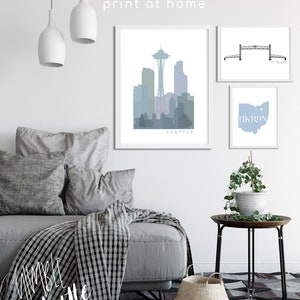 May include: Three printable wall art prints featuring a skyline of Seattle, a bridge, and the state of Ohio with the word "Akron" on it. The prints are framed in white and are displayed on a white wall with a gray couch and a side table with a plant.