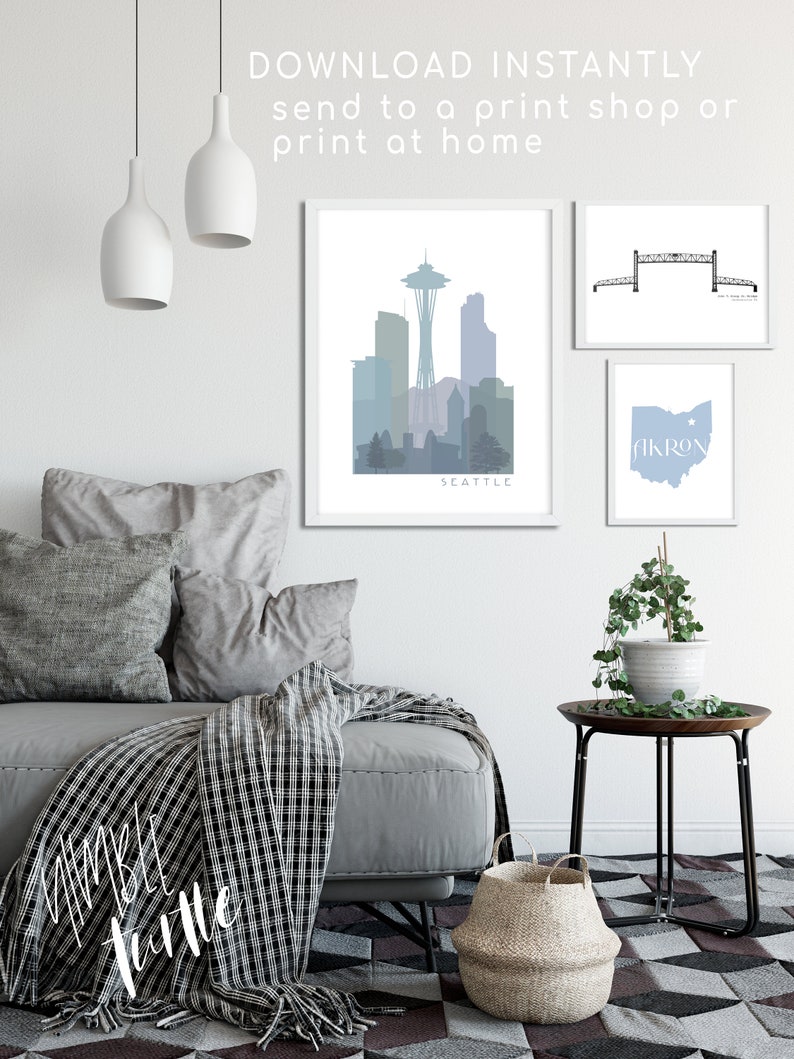 May include: Three printable wall art prints featuring a skyline of Seattle, a bridge, and the state of Ohio with the word "Akron" on it. The prints are framed in white and are displayed on a white wall with a gray couch and a side table with a plant.