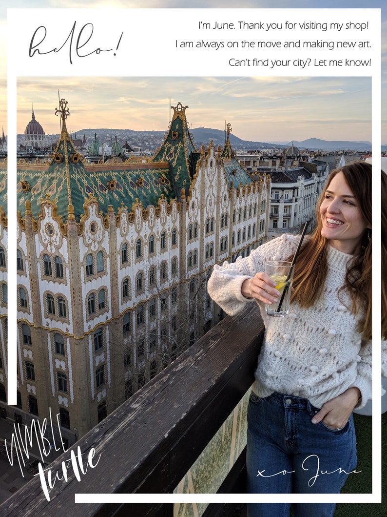 May include: A woman wearing a white sweater and blue jeans stands on a rooftop overlooking a city skyline. She is holding a glass with a lemon wedge and a straw. The text "hello!" is written in black cursive at the top of the image. The text "I'm June. Thank you for visiting my shop! I am always on the move and making new art. Can't find your city? Let me know!" is written in black text below the image. The text "whimble turtle" is written in black cursive at the bottom left of the image. The text "xo June" is written in black cursive at the bottom right of the image.