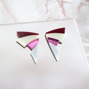 May include: Geometric earrings with a layered design. The earrings feature a combination of colors, including burgundy, cream, light pink, and white. The earrings are made of a reflective material and have a modern, abstract shape.